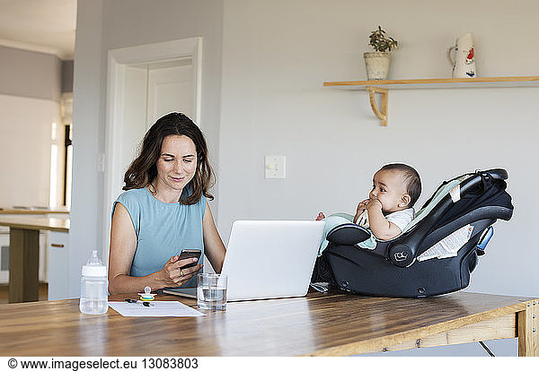 Säugling sitzt im Sitzen und sieht die Mutter an  die ein Smartphone benutzt