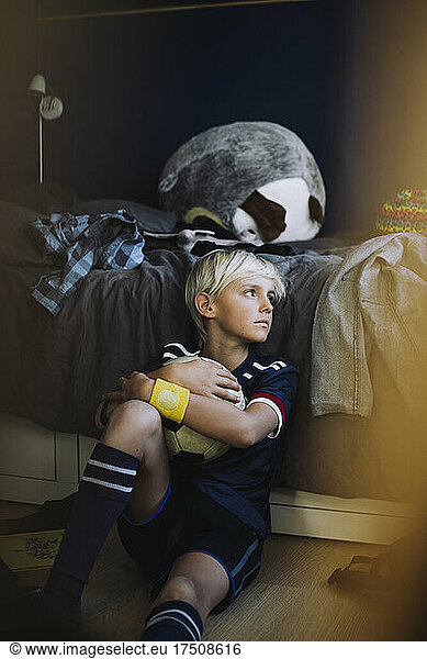 Sad boy with soccer ball looking away in bedroom