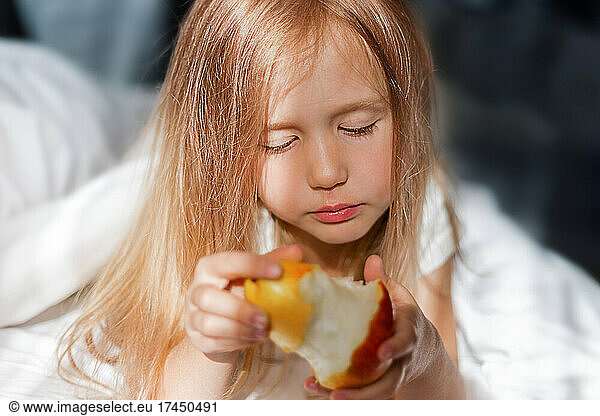 Sad girl in sunbeams eating pear in bed