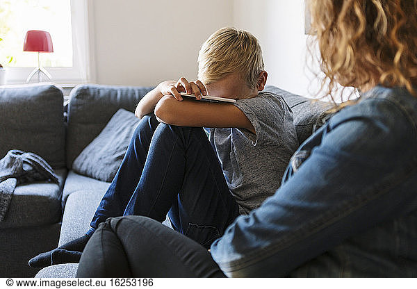 Sad son sitting with mother on sofa at home