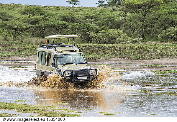 Safari-Fahrzeug spritzt durch eine überschwemmte Schlucht im Ndutu-Gebiet des Ngorongoro-Schutzgebiets in den Serengeti-Ebenen; Tansania