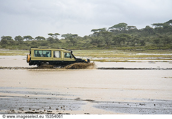 Safari-Fahrzeug spritzt durch eine überschwemmte Schlucht im Ndutu-Gebiet des Ngorongoro-Schutzgebiets in den Serengeti-Ebenen; Tansania