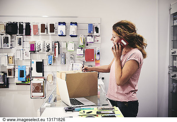 Saleswoman talking on mobile phone while writing on box at counter in electronics store