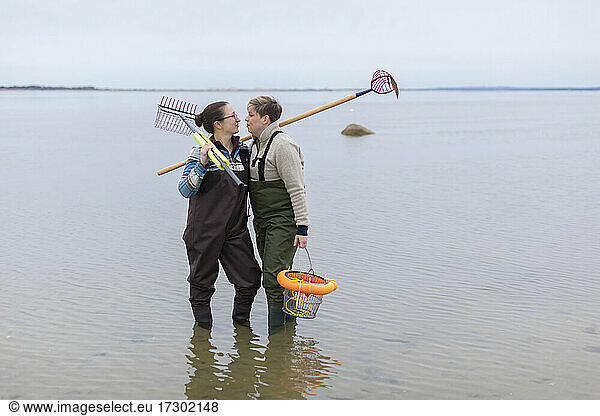 Same sex female couple in waders standing in ocean holding clam rakes