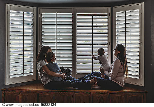 Same sex parents lounging with kids on window seat with tall shutters