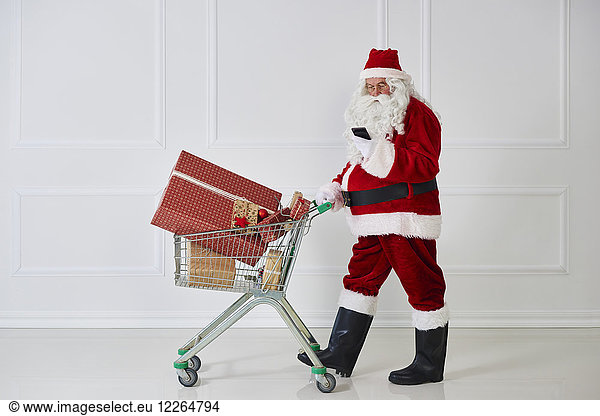 Santa Claus carrying Christmas presents in a shopping cart while looking at cell phone