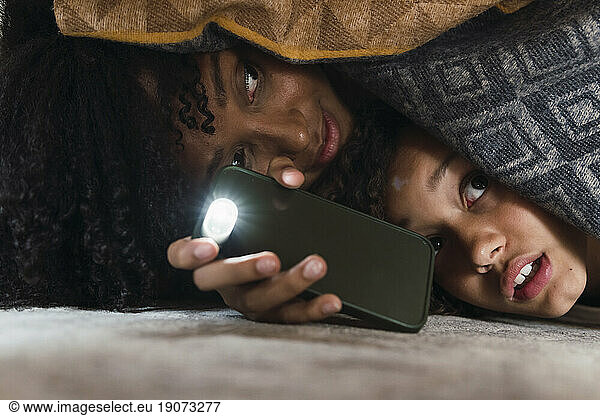 Scared mother and daughter searching floor under couch with smartphone torch