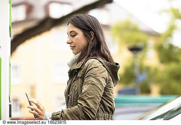 Schöne Frau  die ein Smartphone benutzt  um ein Elektroauto an einer Ladestation zu mieten