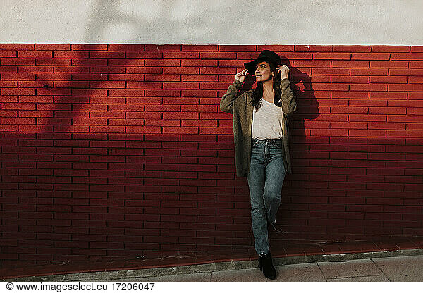 Schöne Frau in Strickjacke und Hut lehnt sich an eine rote Backsteinmauer und schaut weg
