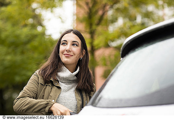 Schöne Frau lädt gemietetes Elektroauto auf  während sie lächelt und wegschaut