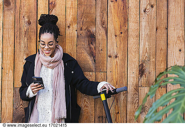 Schöne weibliche Millennial mit Smartphone durch elektrische Push-Roller gegen Holzwand