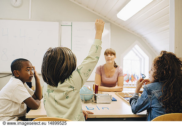 Schoolboy raising hand while answering teacher in classroom at school