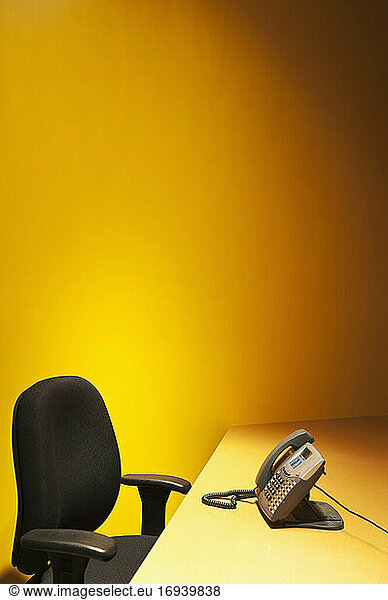 Schreibtisch mit Stuhl und Telefon und gelber Wand dahinter.