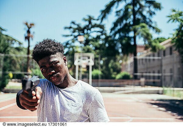 Schwarzer afroamerikanischer Junge auf einem Basketballplatz  der eine Waffe auf die Kamera richtet