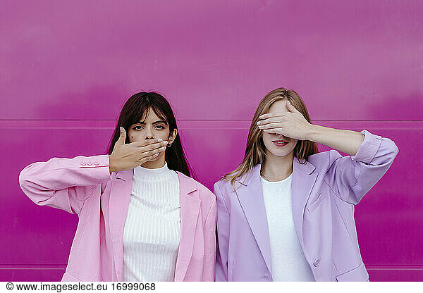 Schwester mit Hand  die den Mund bedeckt  steht neben einer Frau  die die Augen vor einer rosa Wand verdeckt