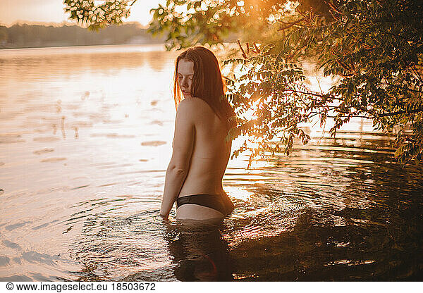 Seductive topless woman standing in lake at sunset