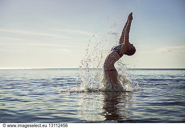 Seductive woman splashing water while diving into sea against sky