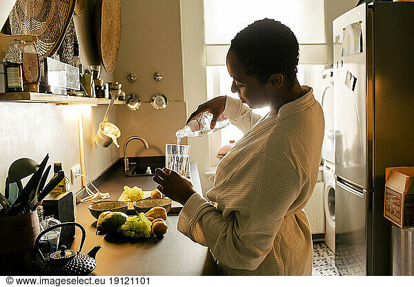 Seitenansicht einer Frau im Bademantel  die am Küchentisch Wasser in ein Glas gießt