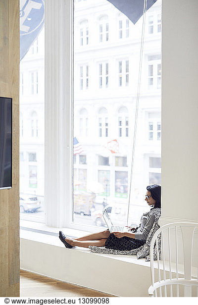 Seitenansicht einer Geschäftsfrau  die einen Laptop benutzt  während sie im Büro auf dem Fensterbrett sitzt