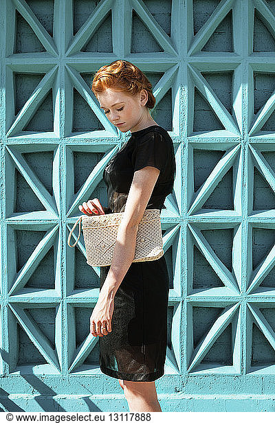 Seitenansicht einer jungen Frau  die eine Clutchbag gegen eine blaue Wand trägt