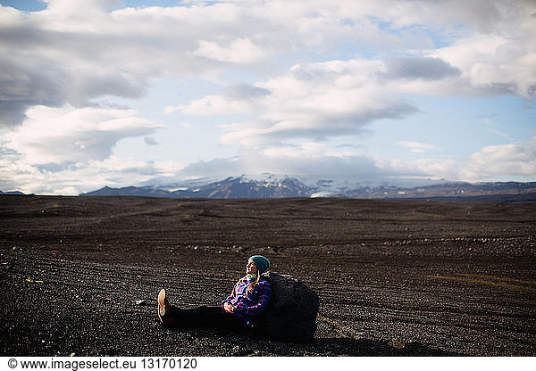Seitenansicht einer mittelgroßen erwachsenen Frau  die an einen Felsbrocken gelehnt auf einer vulkanischen Landschaft sitzt und wegschaut  Island