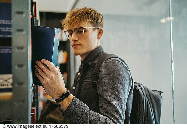 Seitenansicht eines jungen Mannes mit Brille  der ein Buch in der Bibliothek einer Universität durchsucht