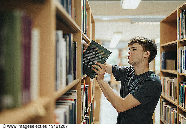 Seitenansicht eines männlichen Studenten  der ein Buch aus einem Bücherregal in der Bibliothek einer Universität entnimmt