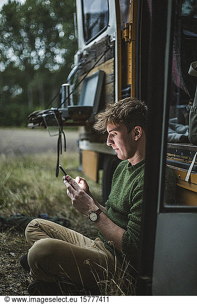 Seitenansicht eines Mannes  der im Schneidersitz sitzt  während er ein Smartphone gegen ein Wohnmobil beim Camping benutzt