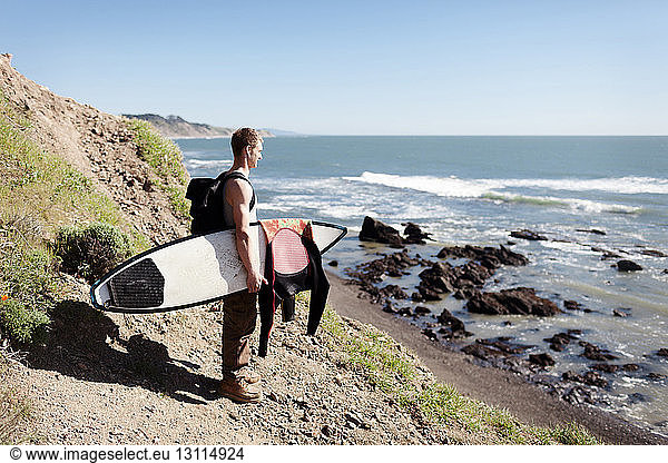 Seitenansicht eines Mannes mit Surfbrett,  der auf einem Berg am Meer vor klarem Himmel steht