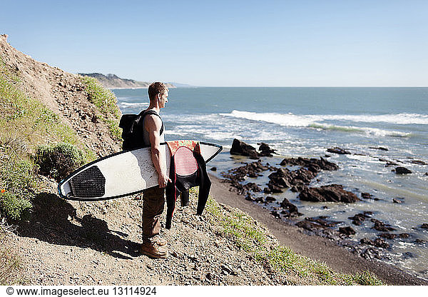 Seitenansicht eines Mannes mit Surfbrett der auf einem Berg am Meer vor klarem Himmel steht