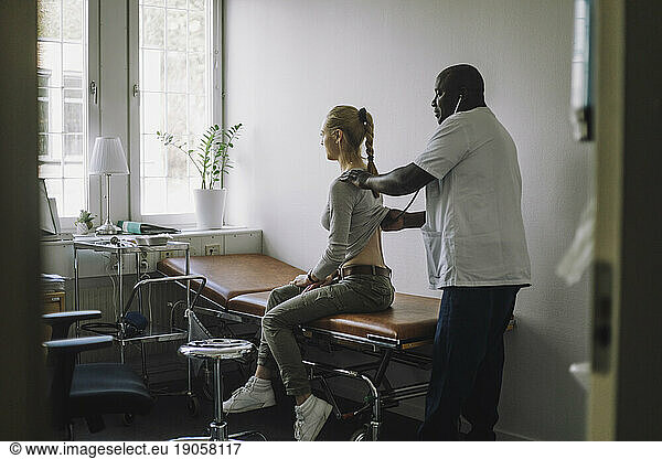 Seitenansicht eines reifen männlichen Arztes  der eine Patientin untersucht  die auf einem Bett in einer Klinik sitzt