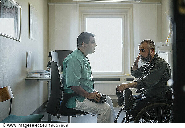 Seitenansicht eines reifen männlichen Arztes  der mit einem im Rollstuhl sitzenden Patienten in einer Klinik spricht