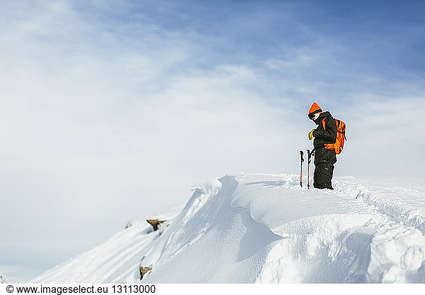 Seitenansicht eines Wanderers mit Rucksack und Skistöcken  der auf einem schneebedeckten Berg vor bewölktem Himmel steht