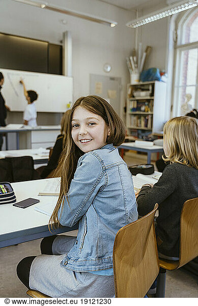 Seitenansicht Porträt eines lächelnden Mädchens  das eine Jeansjacke trägt und auf einem Stuhl im Klassenzimmer sitzt