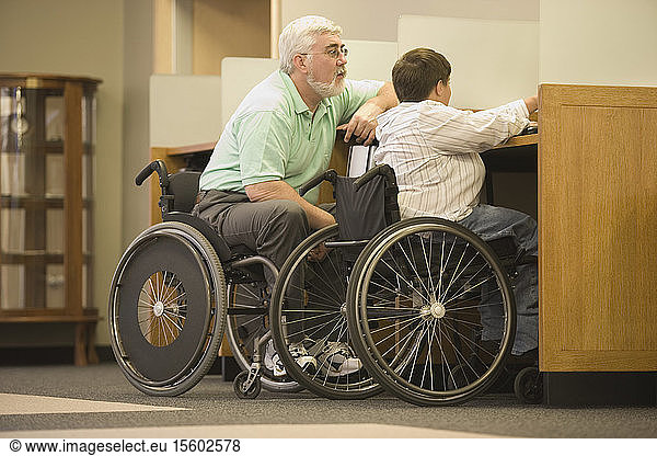 Seitenprofil eines Mannes mittleren Alters und einer Frau mittleren Alters mit Muskeldystrophie  die in einer Bibliothek in Rollstühlen sitzen
