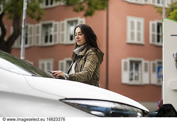 Selbstbewusste schöne Frau  die ein Smartphone benutzt  um ein Elektroauto an einer Ladestation zu mieten