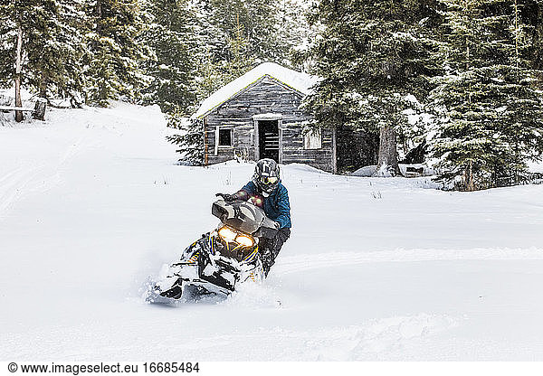 selbstbewusster Mann auf Schneemobil mit alter Hütte dahinter.