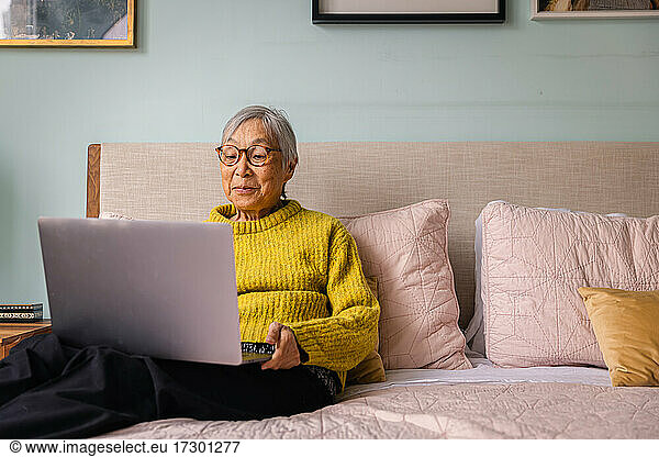 Senior asian woman using laptop while sitting in bedroom at home