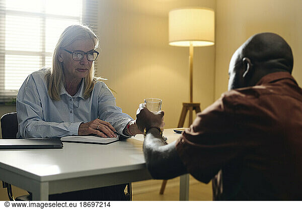 Senior blond psychologist giving glass of water to patient at office