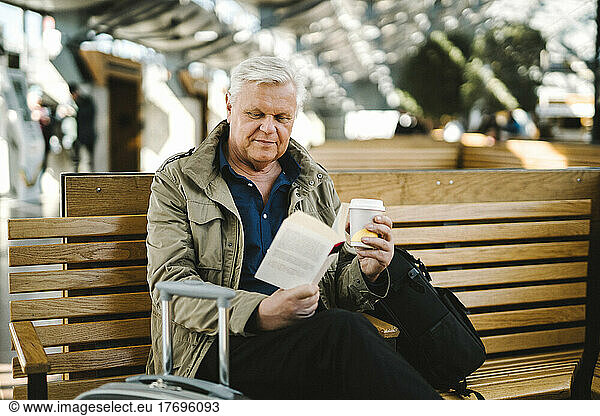 Senior businessman holding disposable cup reading book while sitting on bench at railroad station food court