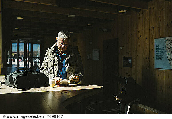 Senior businessman reading book while standing in sunlight at railroad station waiting room
