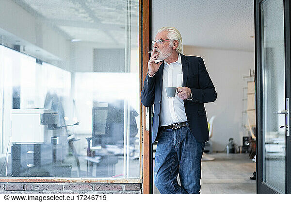 Senior businessman smoking cigarette while leaning on glass wall at office