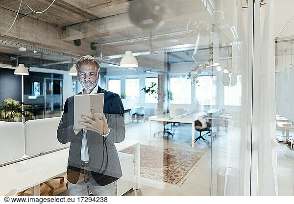 Senior businessman using digital tablet while standing behind glass wall
