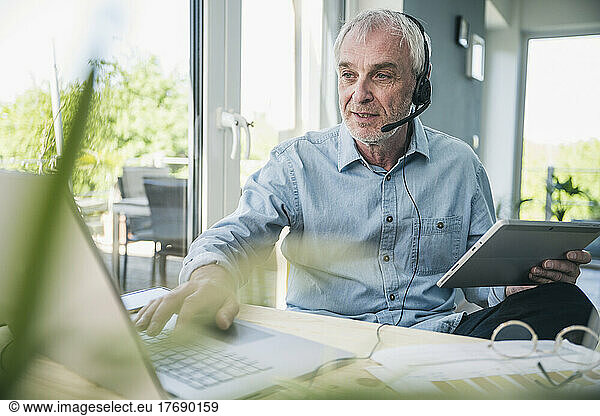 Senior businessman wearing headset working on laptop at home