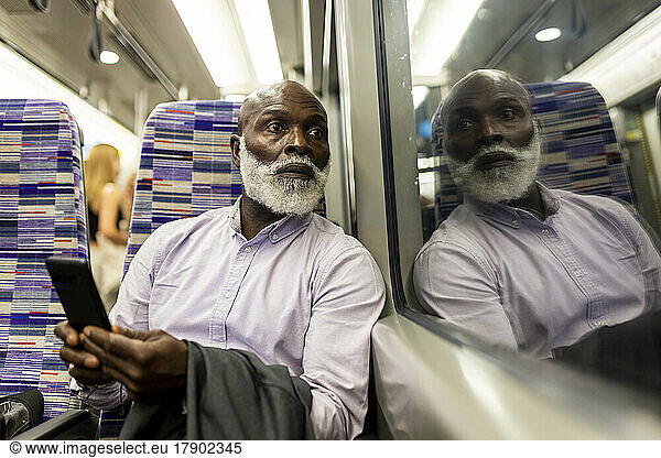 Senior businessman with smart phone looking through window in train