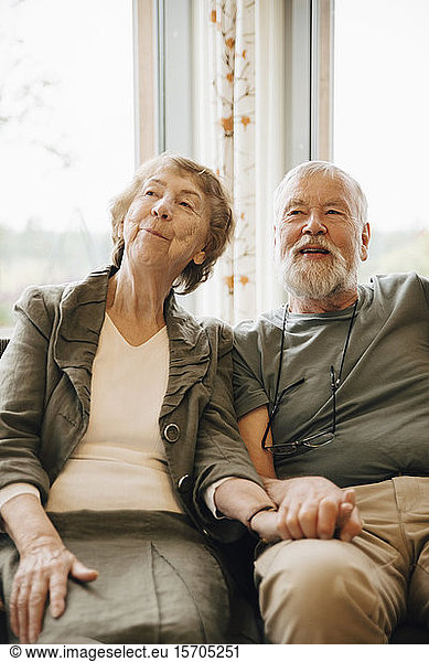Senior couple holding hands while sitting against window at nursing home