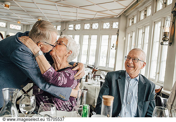 Senior couple kissing while male friend smiling in restaurant