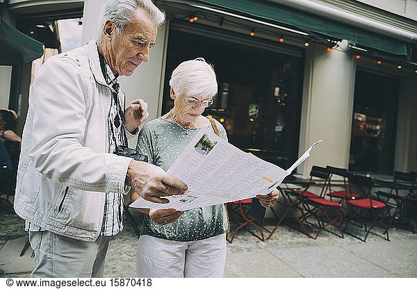 Senior couple reading map while standing on sidewalk in city during vacation