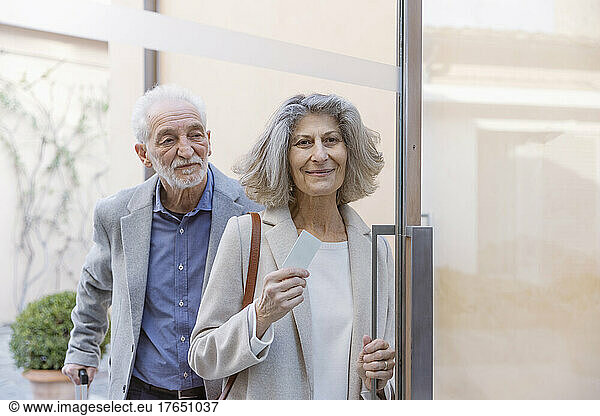 Senior couple with cardkey standing at glass door of boutique hotel