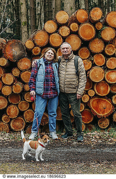 Senior couple with dog standing in front of pine logs in forest